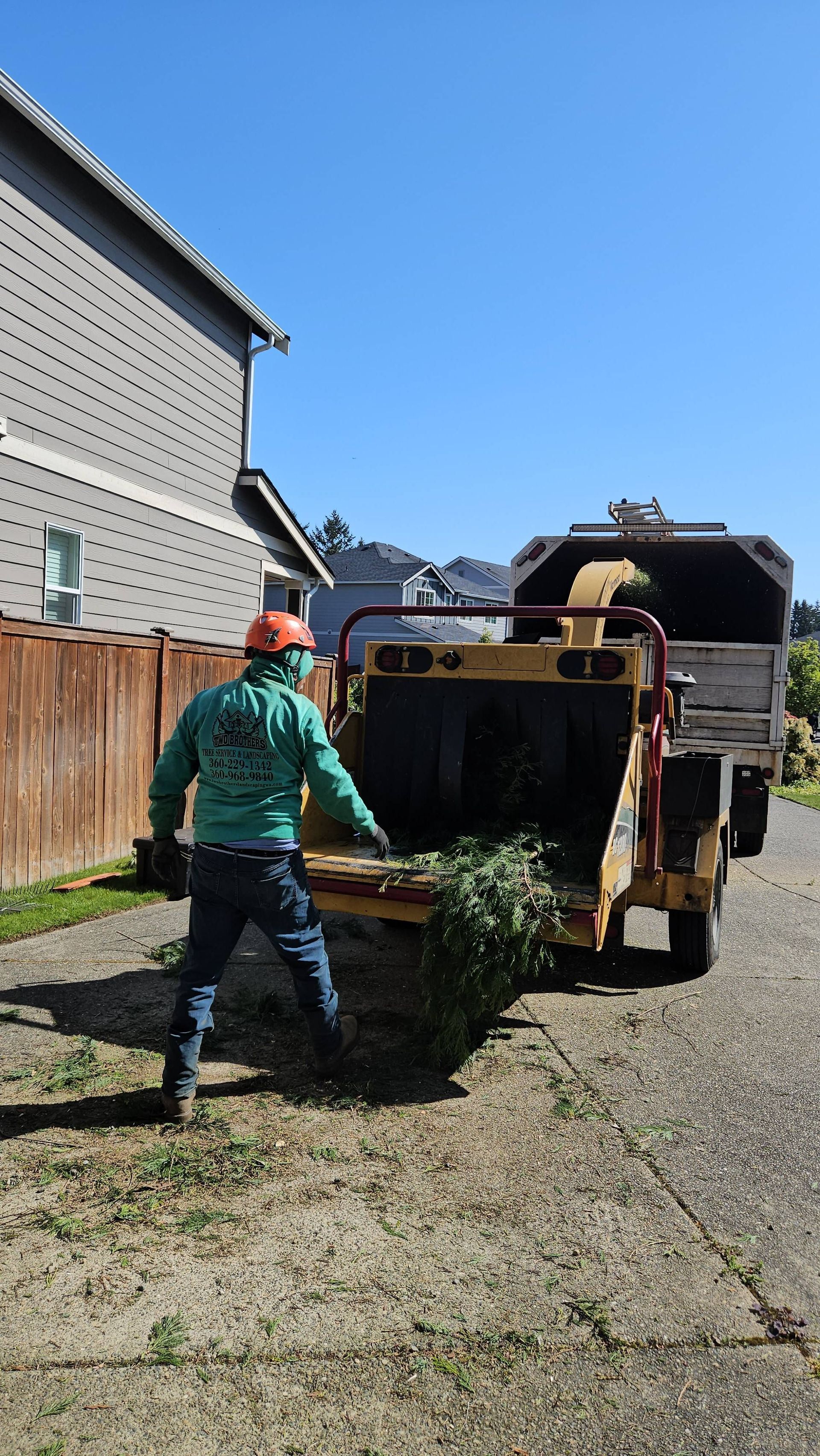 Worker loading debris into a dumpster in a driveway beside a house on a sunny day