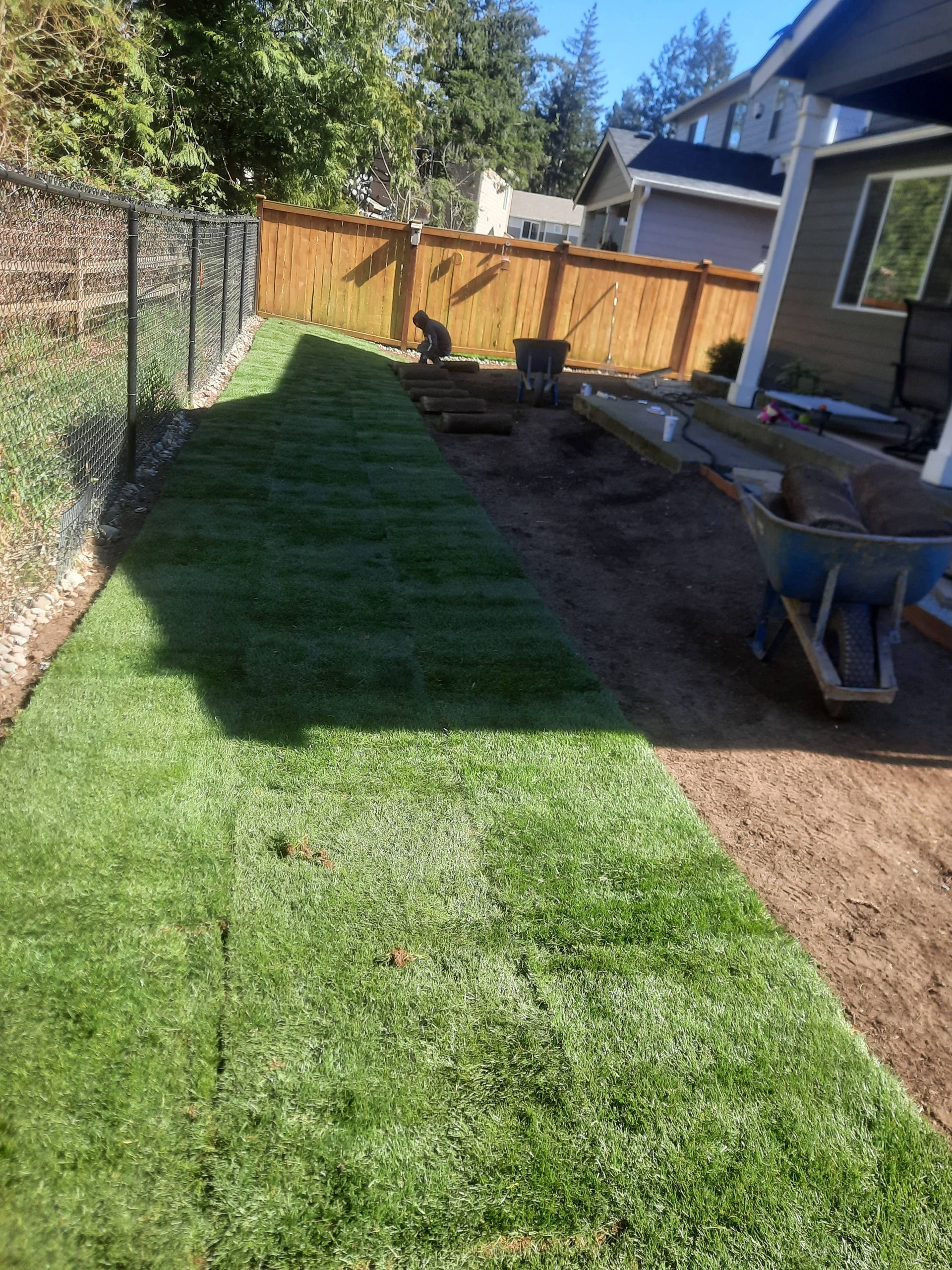 Backyard with a green lawn, dirt strip, wooden fence, and a wheelbarrow beside a house patio.
