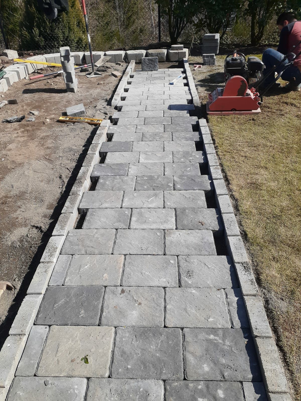 Stone walkway under construction in a yard, with pavers, edging, and tools along the sides.