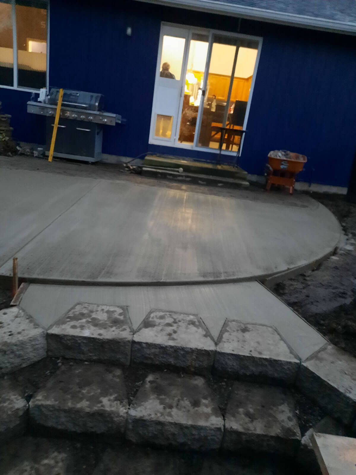 Stone steps lead to a concrete patio in front of a blue building with lit glass doors at night