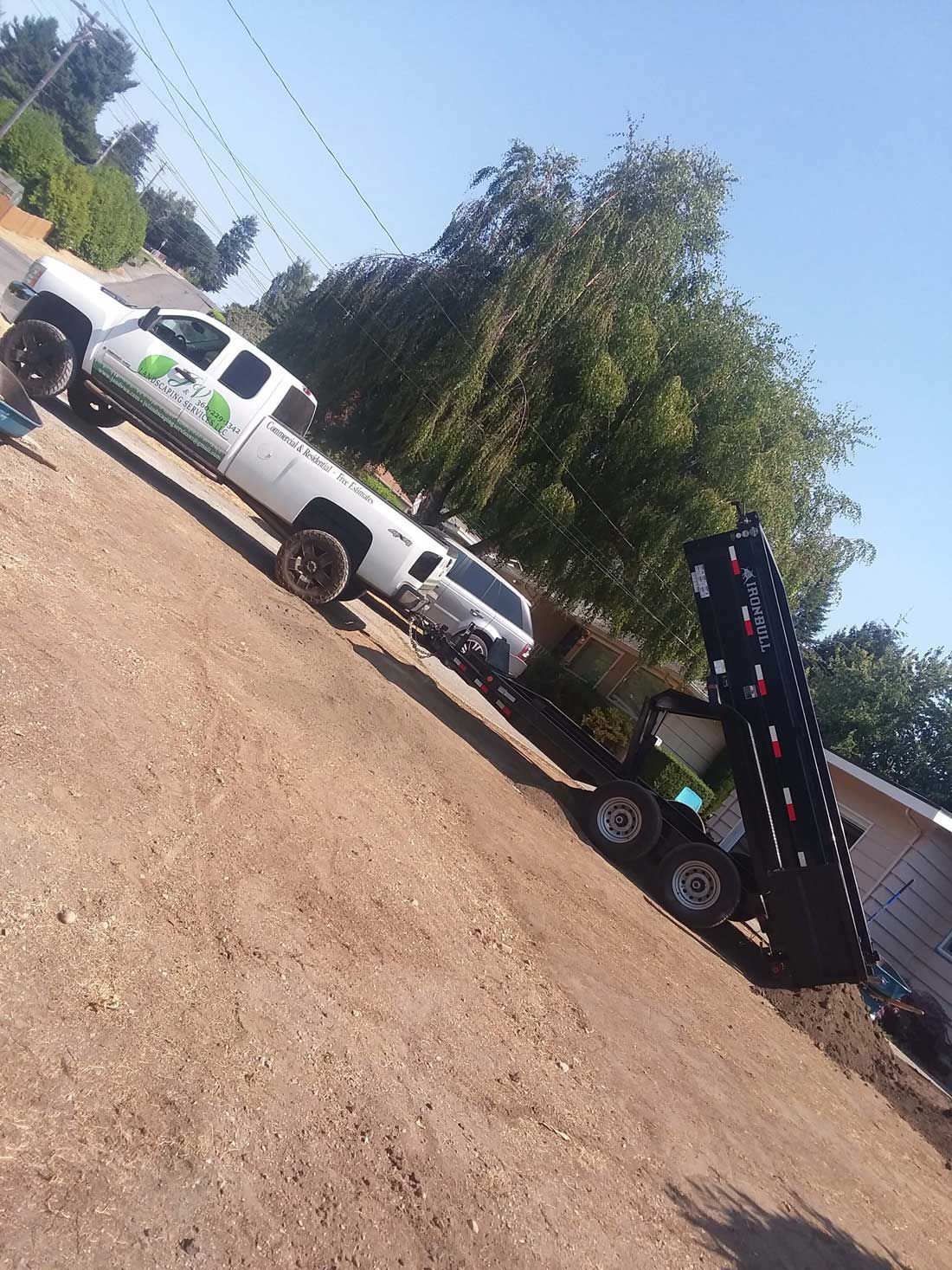 White utility trucks parked on a dirt lot beside a tree and building, with a raised dump bed in the foreground
