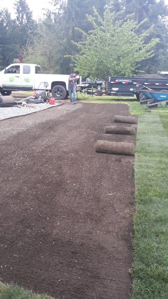 Gravel driveway under construction with rolled sod, parked trucks, and trees in the background