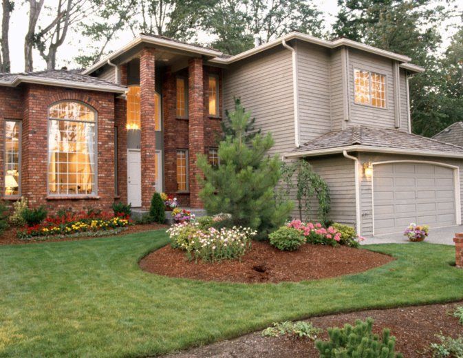 Brick suburban house with lit windows, garage, and landscaped front yard at dusk