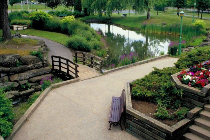 Curved garden path beside a pond, with stone steps, a small bridge, and colorful flower beds.