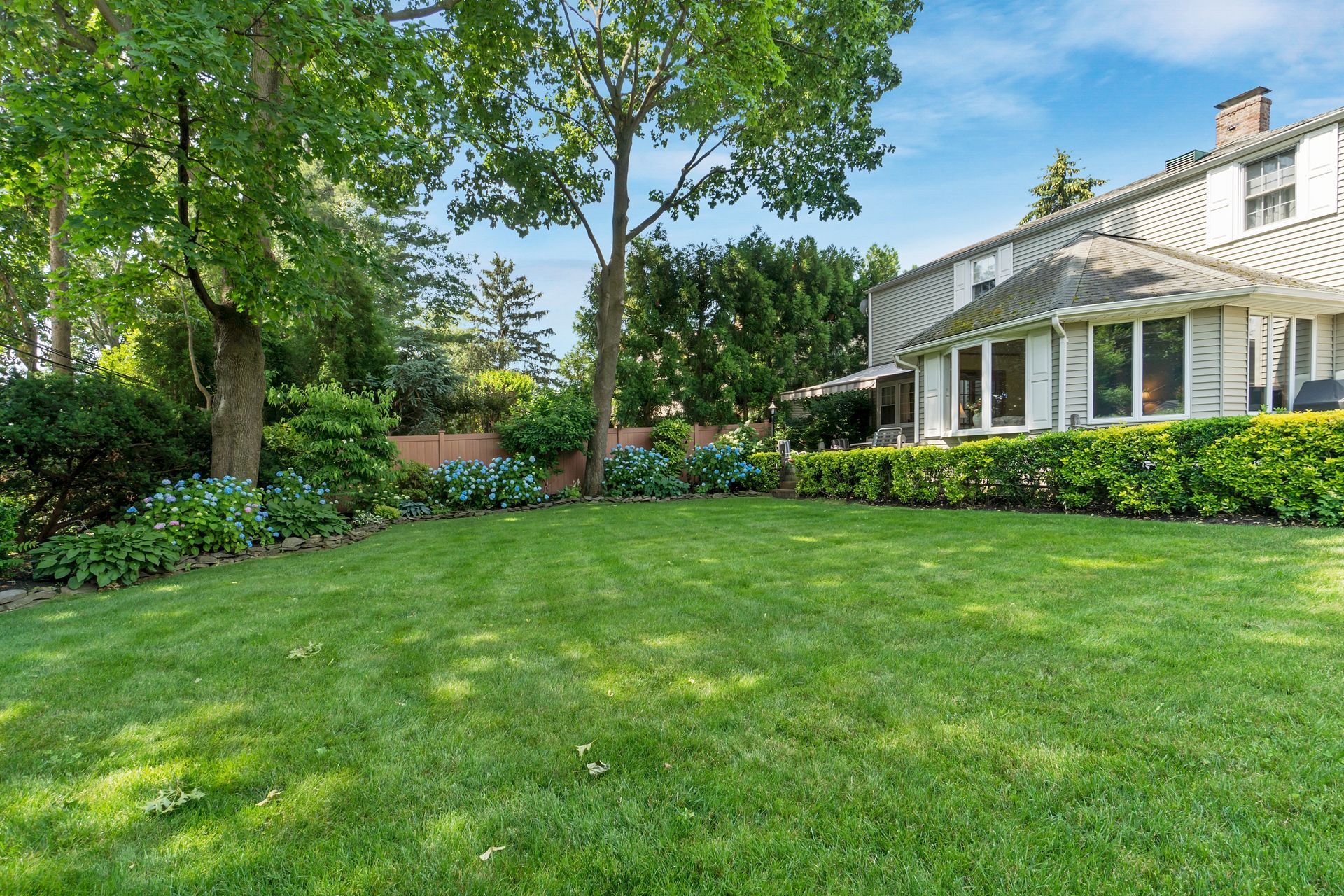 Lush green backyard lawn beside a white house with hedges, trees, and a covered porch