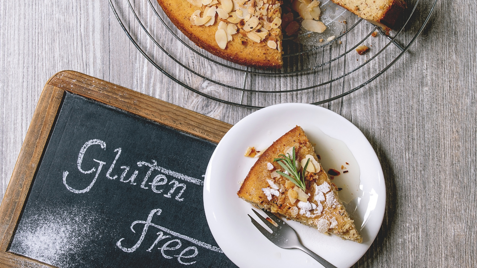 A chalk board with Gluten-Free written on it in chalk. A piece of pie on a white plate with a silver fork. On the pie is a spring of rosemary and powdered sugar.