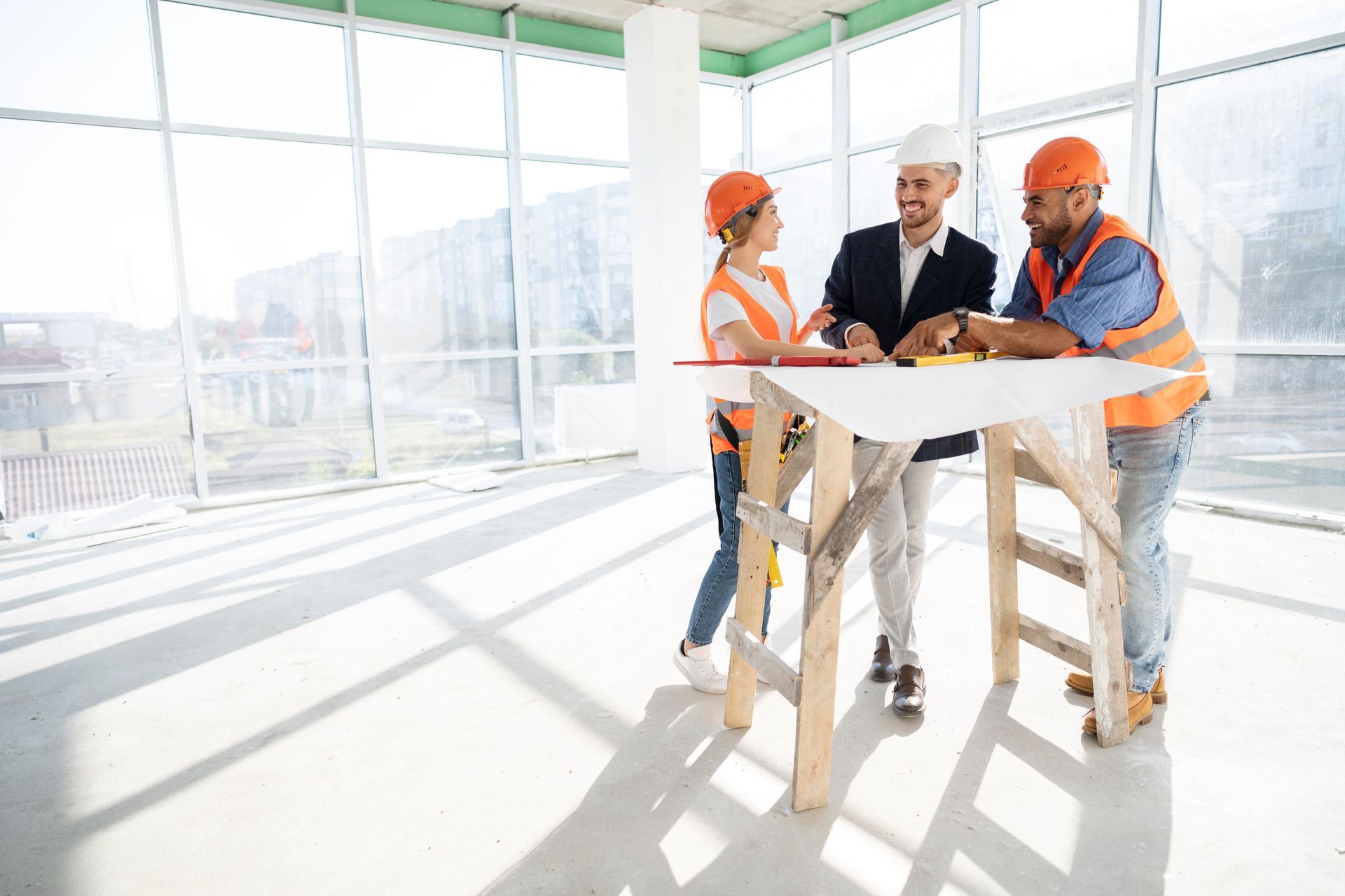 Construction workers and architect discussing plans at a construction site, near large windows; smiling, using blueprint.