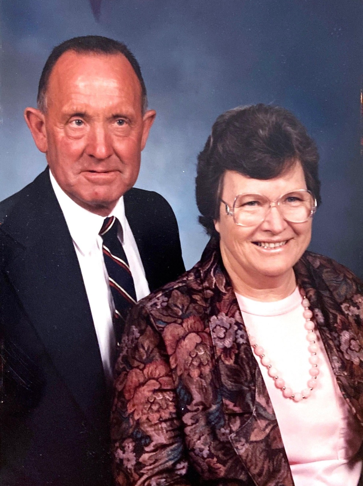 A studio portrait of a man in a suit and tie standing next to a smiling woman wearing a floral blazer and pink necklace.