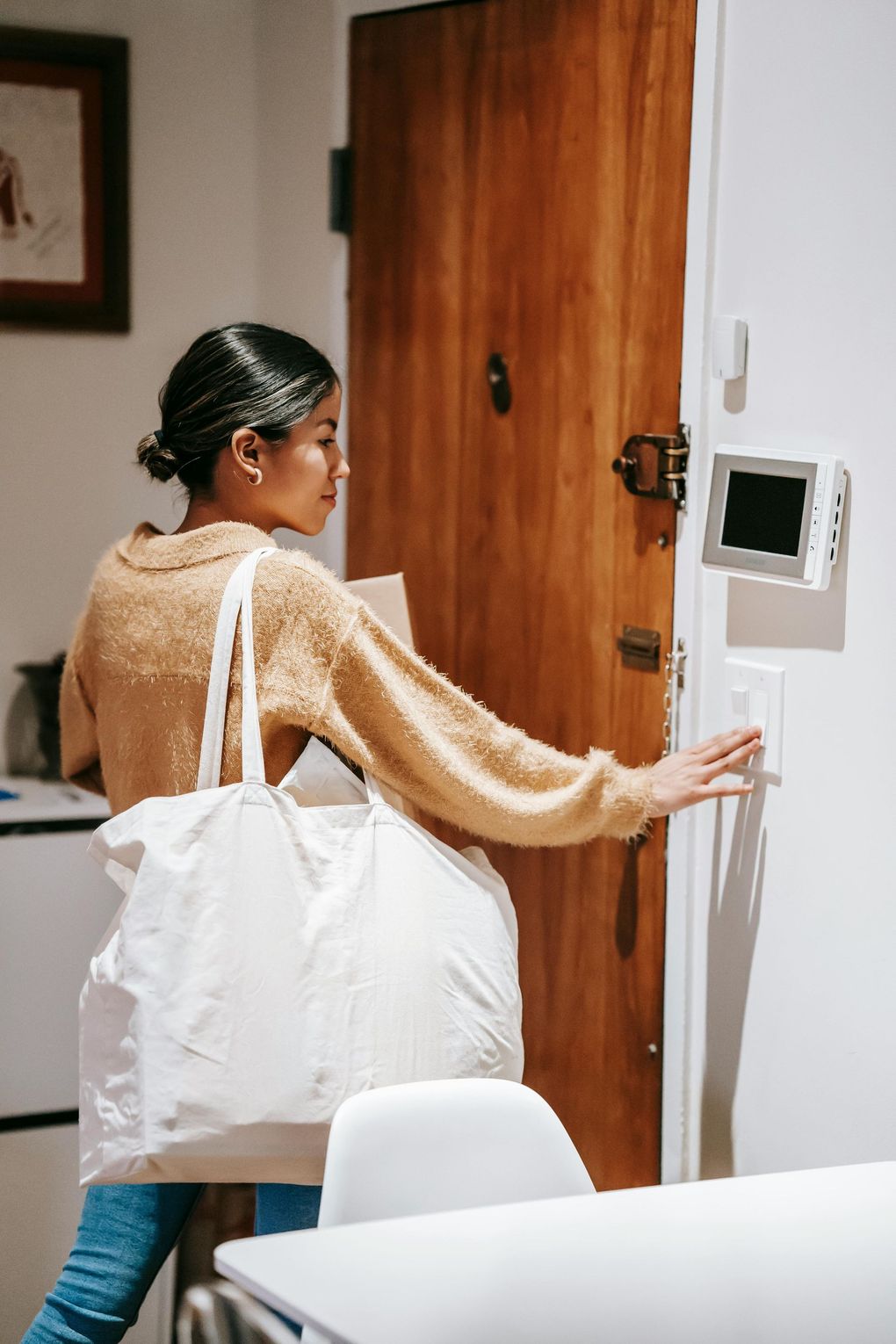 A person wearing a tan sweater and carrying a white tote bag reaches to flip a light switch on a wall near a wooden door.