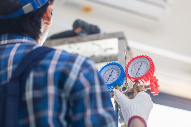 Technician wearing a blue plaid shirt and white gloves holds a set of blue and red HVAC pressure gauges.