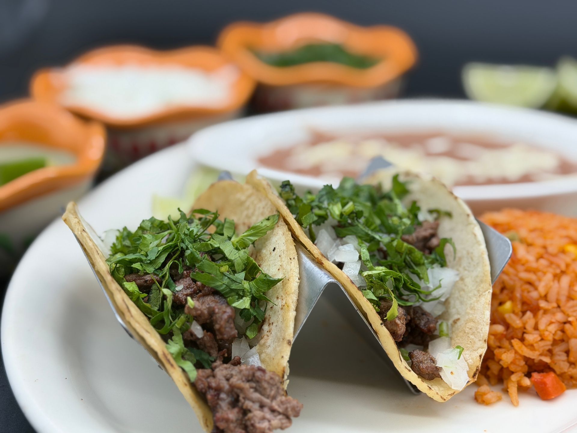 Tacos with chopped cilantro and meat on a plate, with rice and bowls of salsa in the background