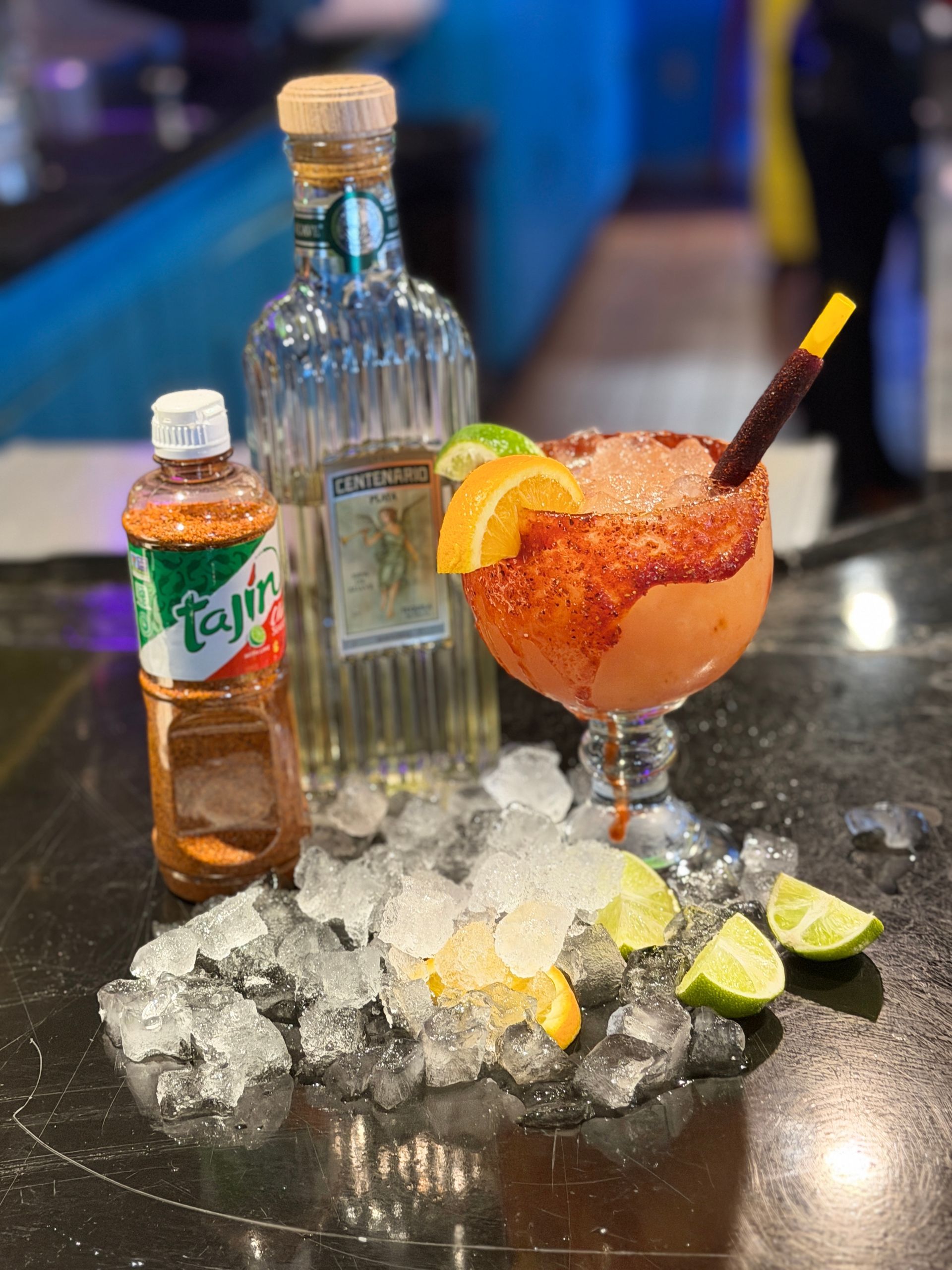 Cocktail on crushed ice with lime wedges, a small drink bottle, and a liquor bottle on a bar counter