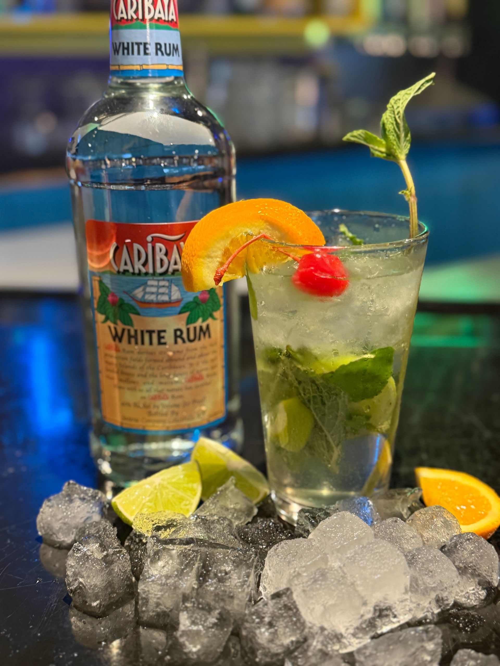 White rum bottle with a mojito cocktail on ice, garnished with mint, lime, orange, and cherry.