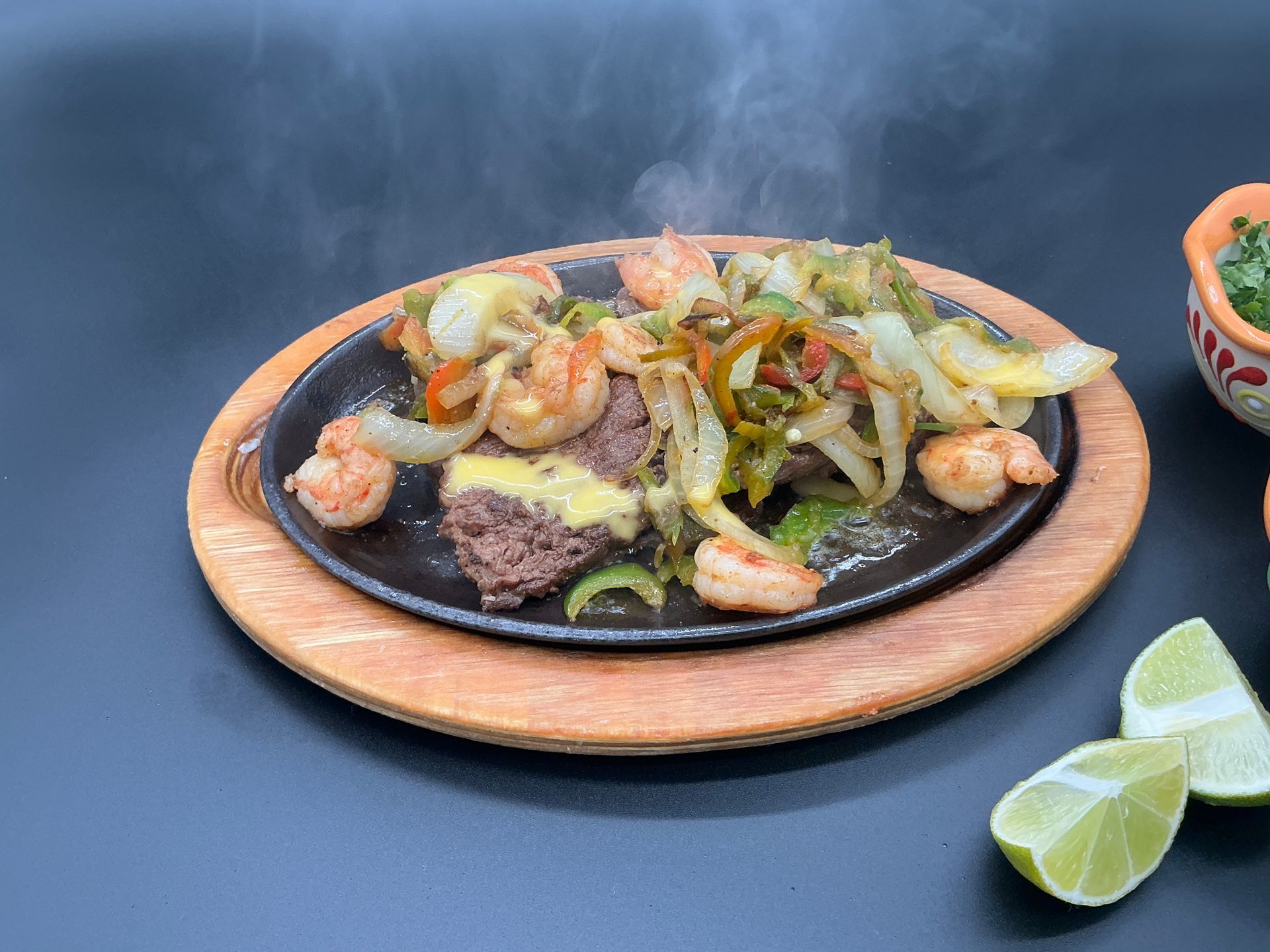 Sizzling fajita platter with shrimp, beef, peppers, onions, and lime wedges on a dark table
