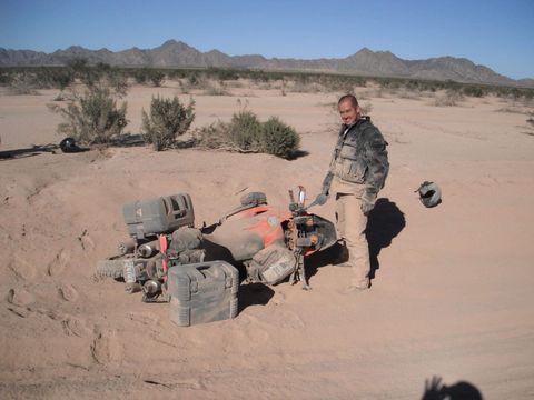 Man stands next to overturned ATV in desert sand. Brown landscape, mountains in the distance.