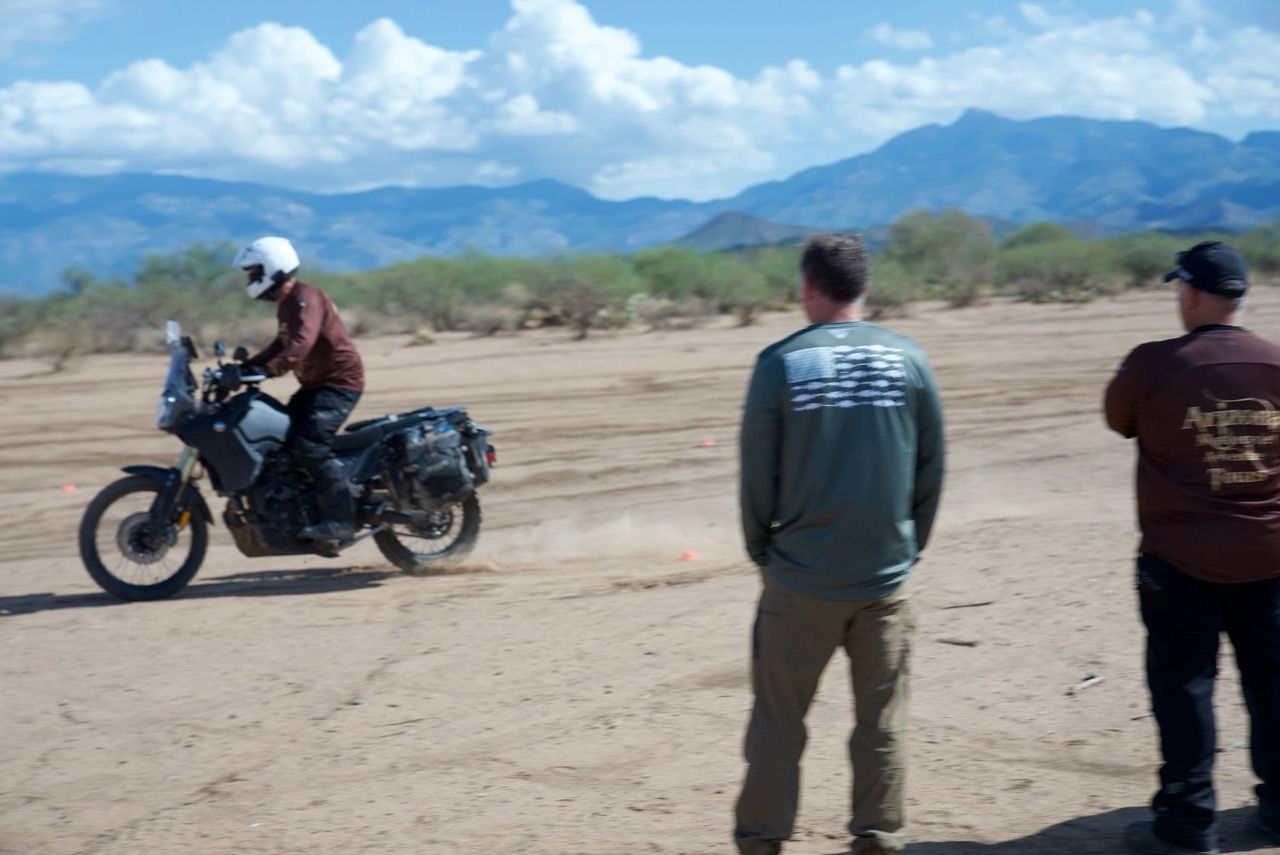 Motorcyclist riding on dirt track, two men watch. Mountains and blue sky in background.