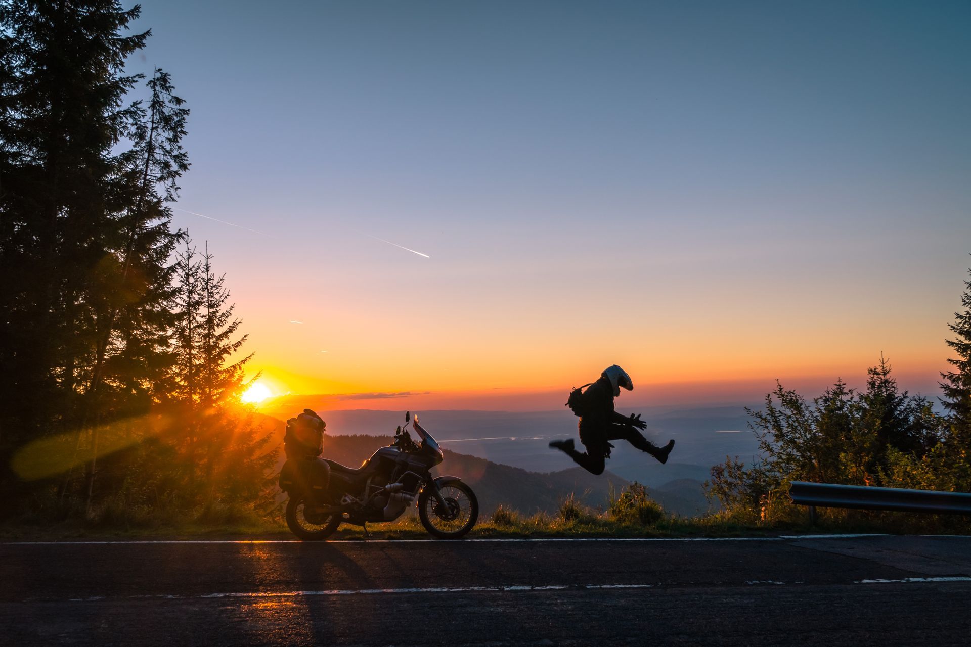 Motorcyclist leaps near his bike on a mountain road at sunset. Orange sky and forest silhouette.