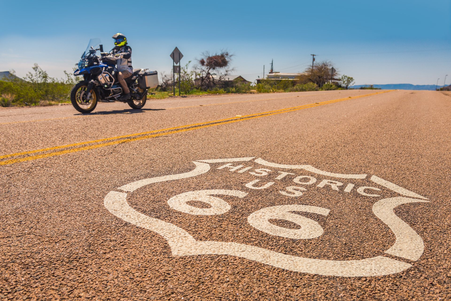 Motorcyclist on Route 66, landmark sign in foreground. Asphalt road, blue sky, and dry landscape.