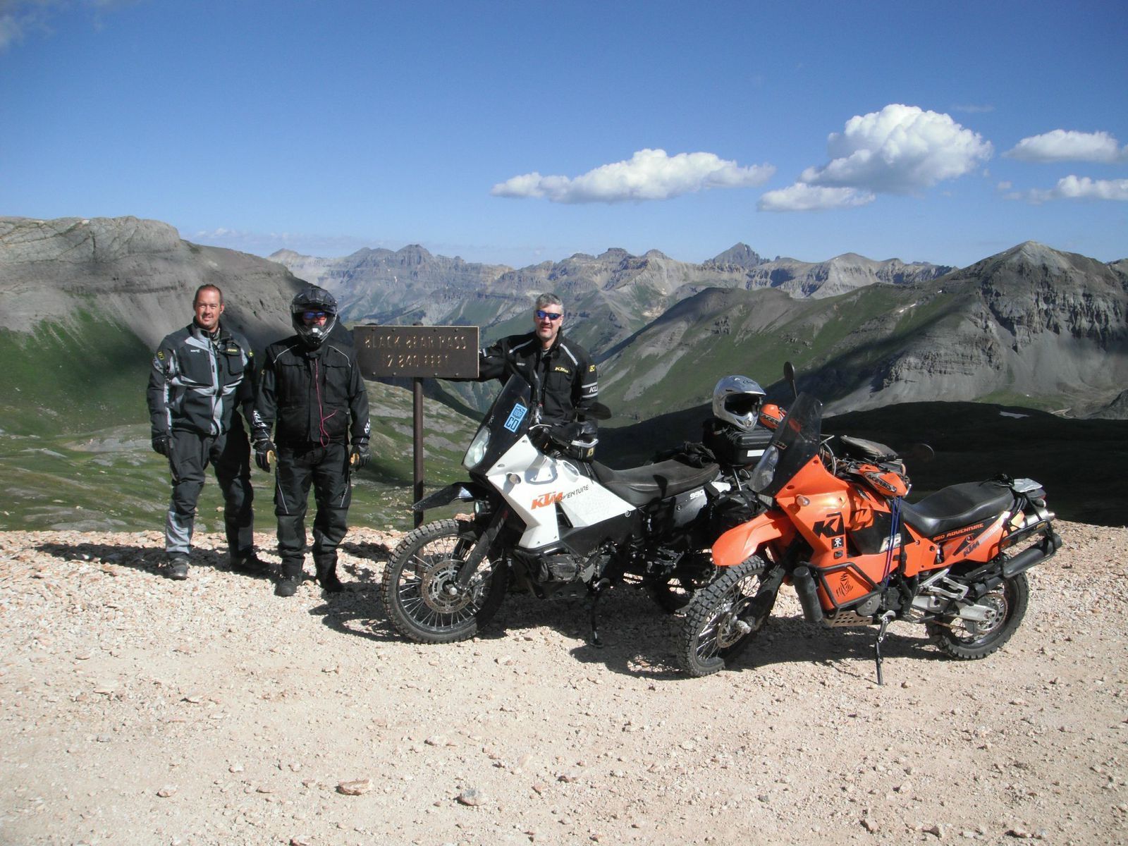 Three people stand with two motorcycles at a mountain overlook under a blue sky.