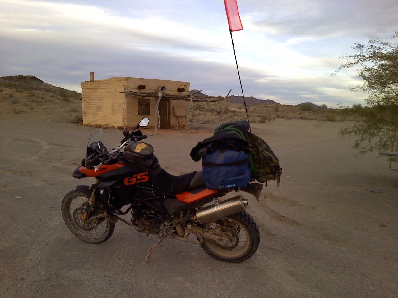 Motorcycle parked near a small building in a desert landscape. Pink flag on a pole, packs on the bike.