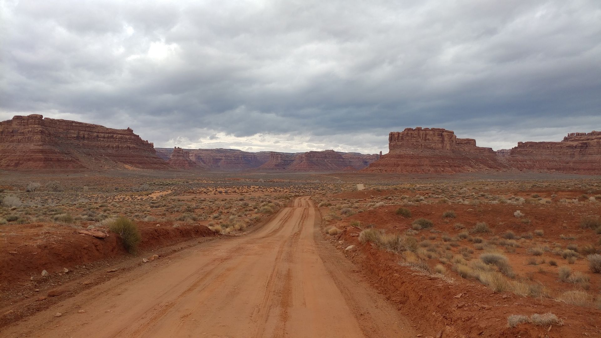 Dirt road through a desert landscape with red rock formations under a cloudy sky.