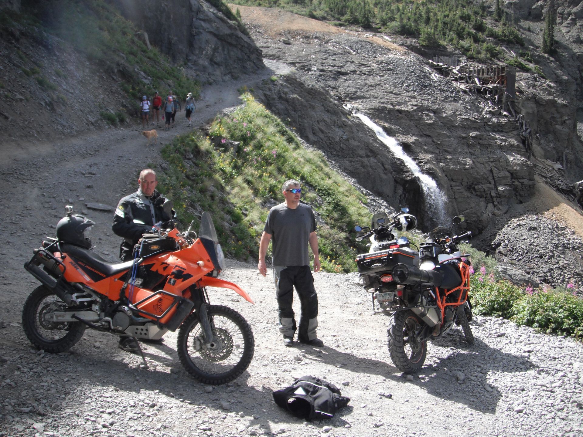 Two motorcycles parked on a rocky mountain road, riders nearby. Waterfall and hikers in background.