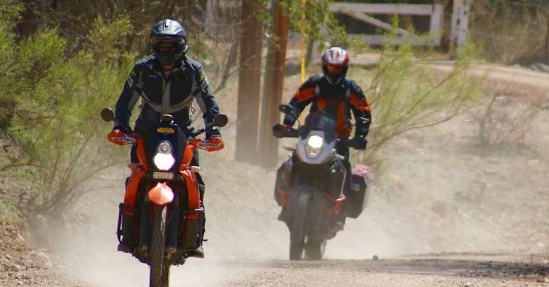 Two motorcyclists riding dirt bikes on a dusty road; one in black gear, the other in orange.