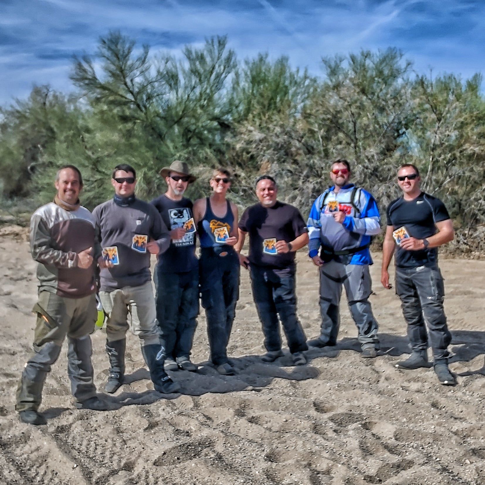 Group of seven people standing on sand, trees in background, holding small items. Sunny, outdoor.
