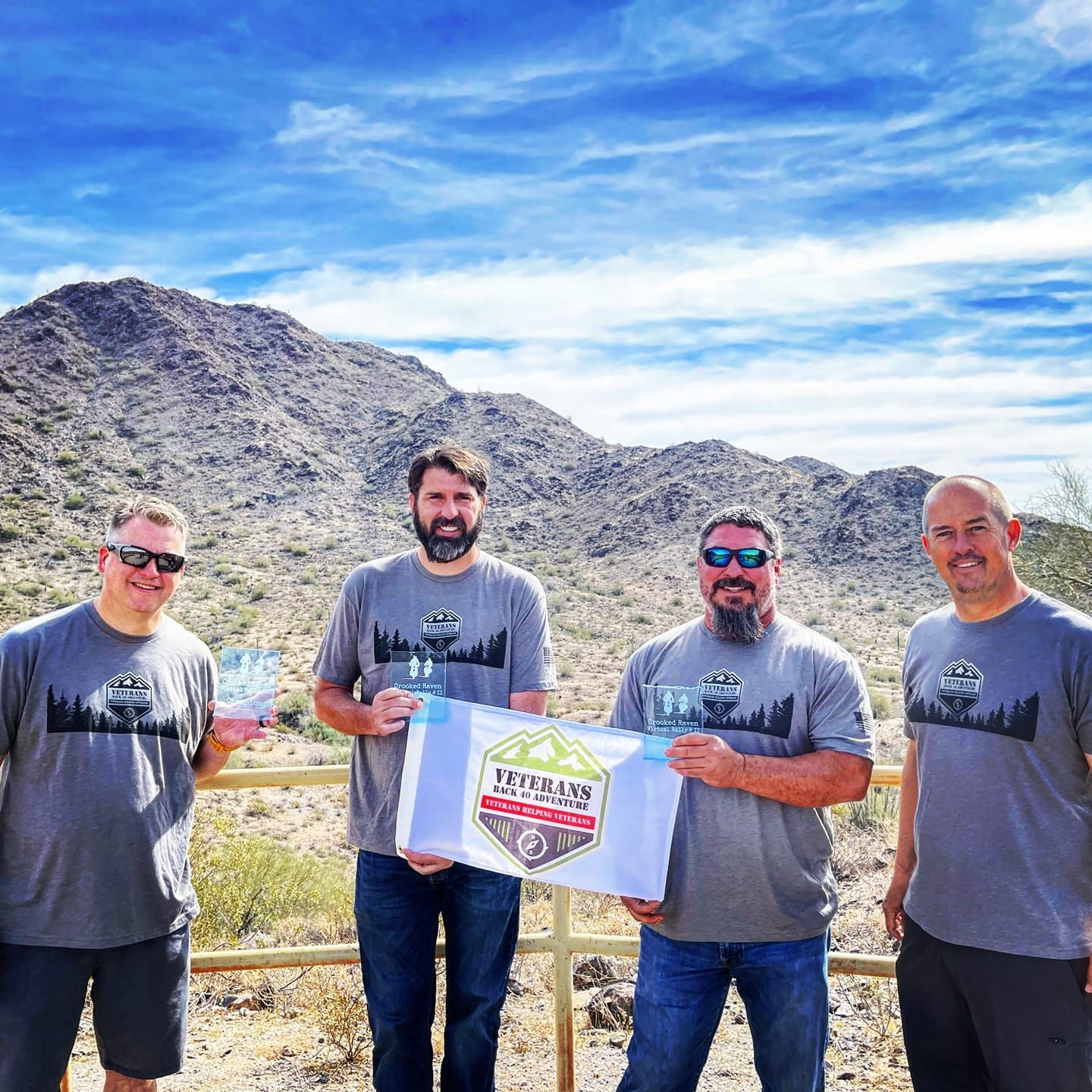 Four men hold awards outdoors with a mountain backdrop, wearing matching gray t-shirts.