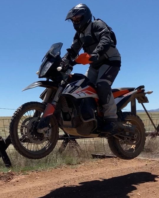 Motorcyclist on an orange and black adventure bike jumping a low barrier outdoors under a blue sky.
