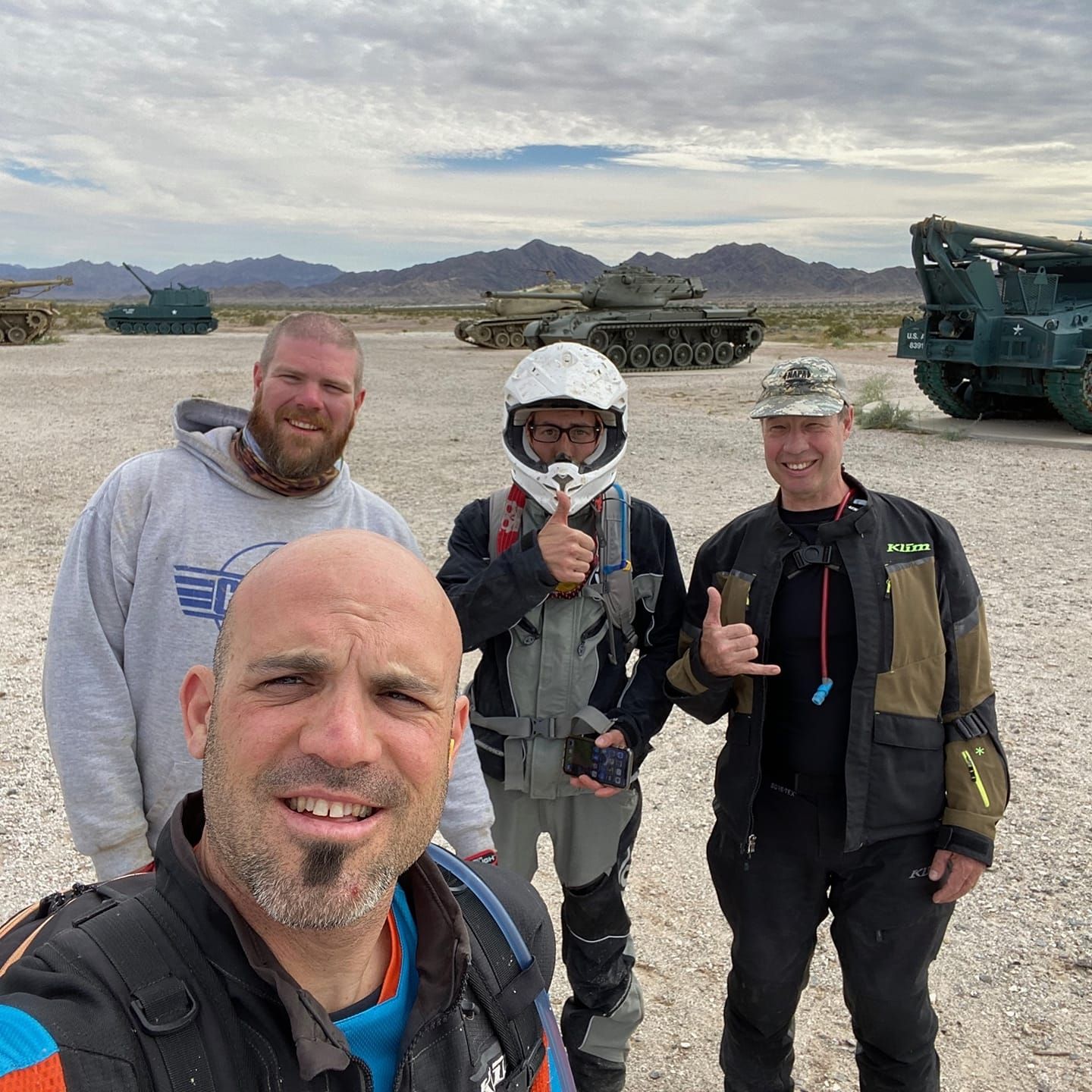Four men posing for a photo near military tanks in a desert landscape; one giving a thumbs up.