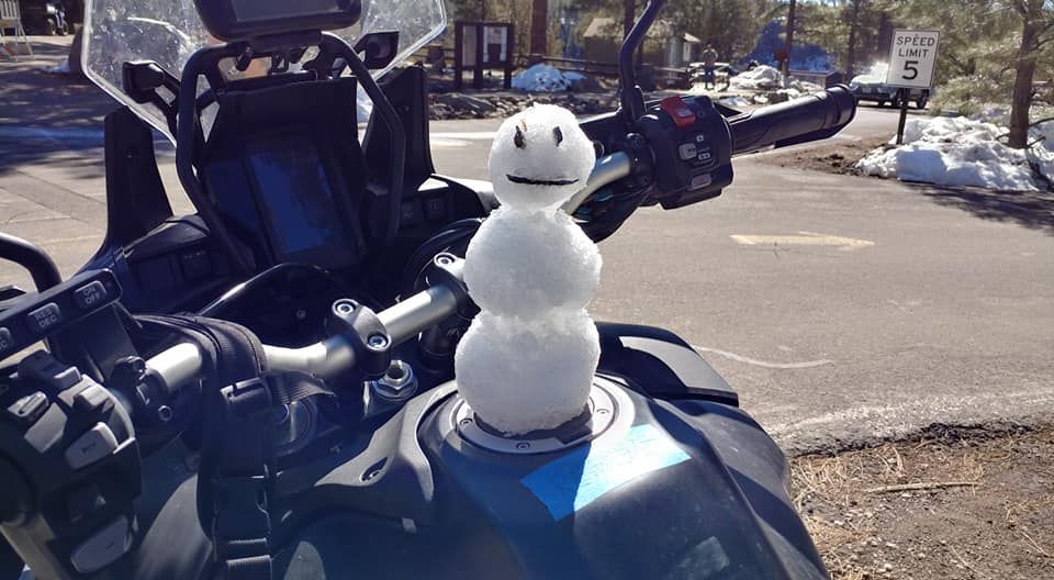 Snowman perched on a motorcycle's fuel tank, smiling. Snowy roadside setting.