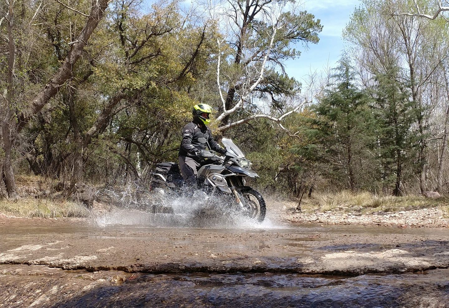 Motorcyclist rides a motorcycle through a water puddle on a dirt road, trees in the background.