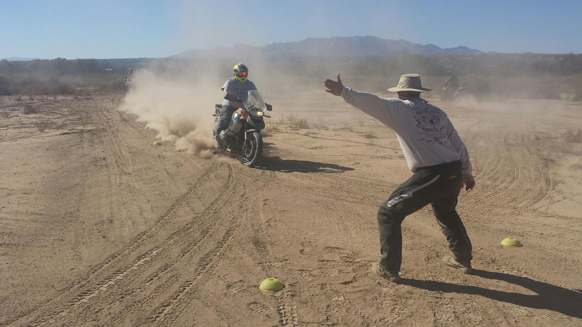 Man gesturing towards a motorcycle kicking up dust on a dirt road. Two yellow balls are on the ground.