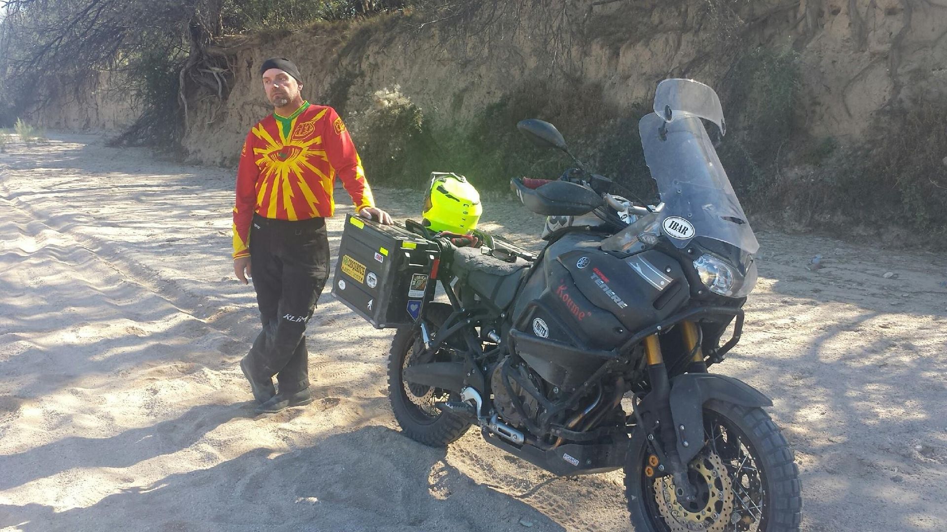 Man with motorcycle on a sandy beach; wearing colorful shirt, standing beside a black adventure bike with gear.