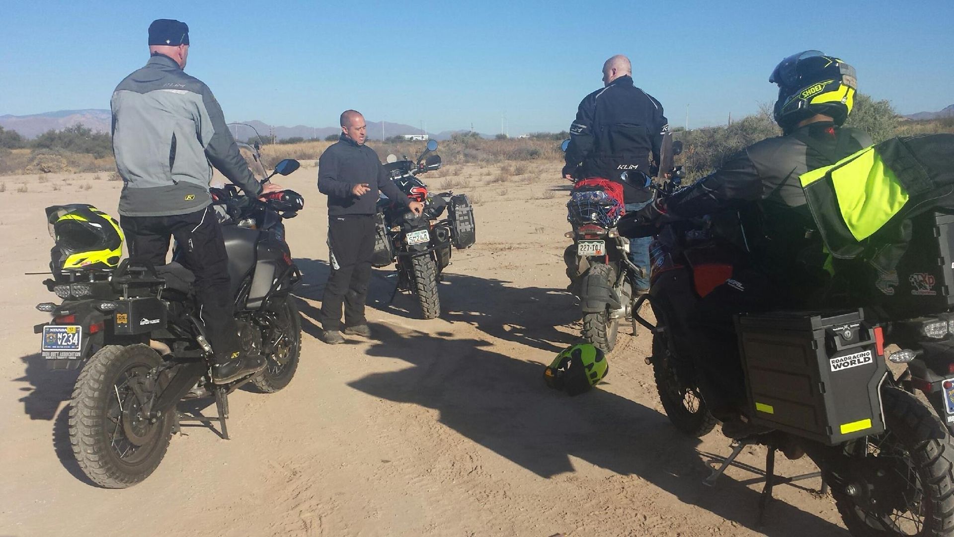 Four motorcyclists on dirt bikes stop on a dirt road in a desert landscape.