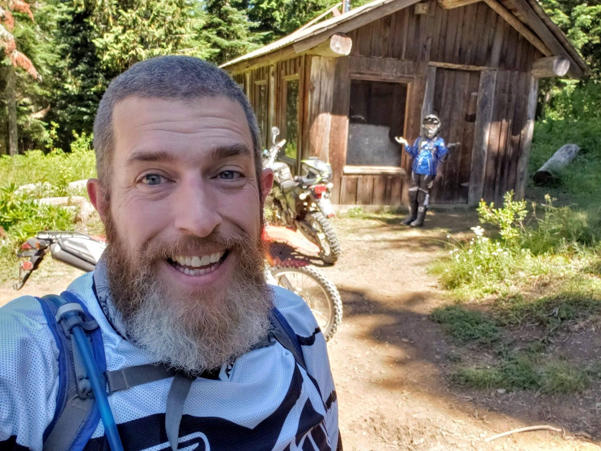Man with beard smiles for selfie with dirt bike. Another person stands by wooden outhouse.