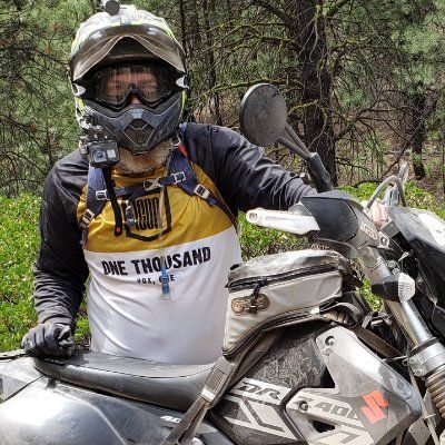 Person in helmet and riding gear on a Suzuki DR400 motorcycle in a wooded area.