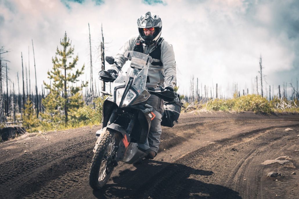 Motorcyclist on an off-road trail, riding a KTM adventure bike. Cloudy sky and burned trees in the background.