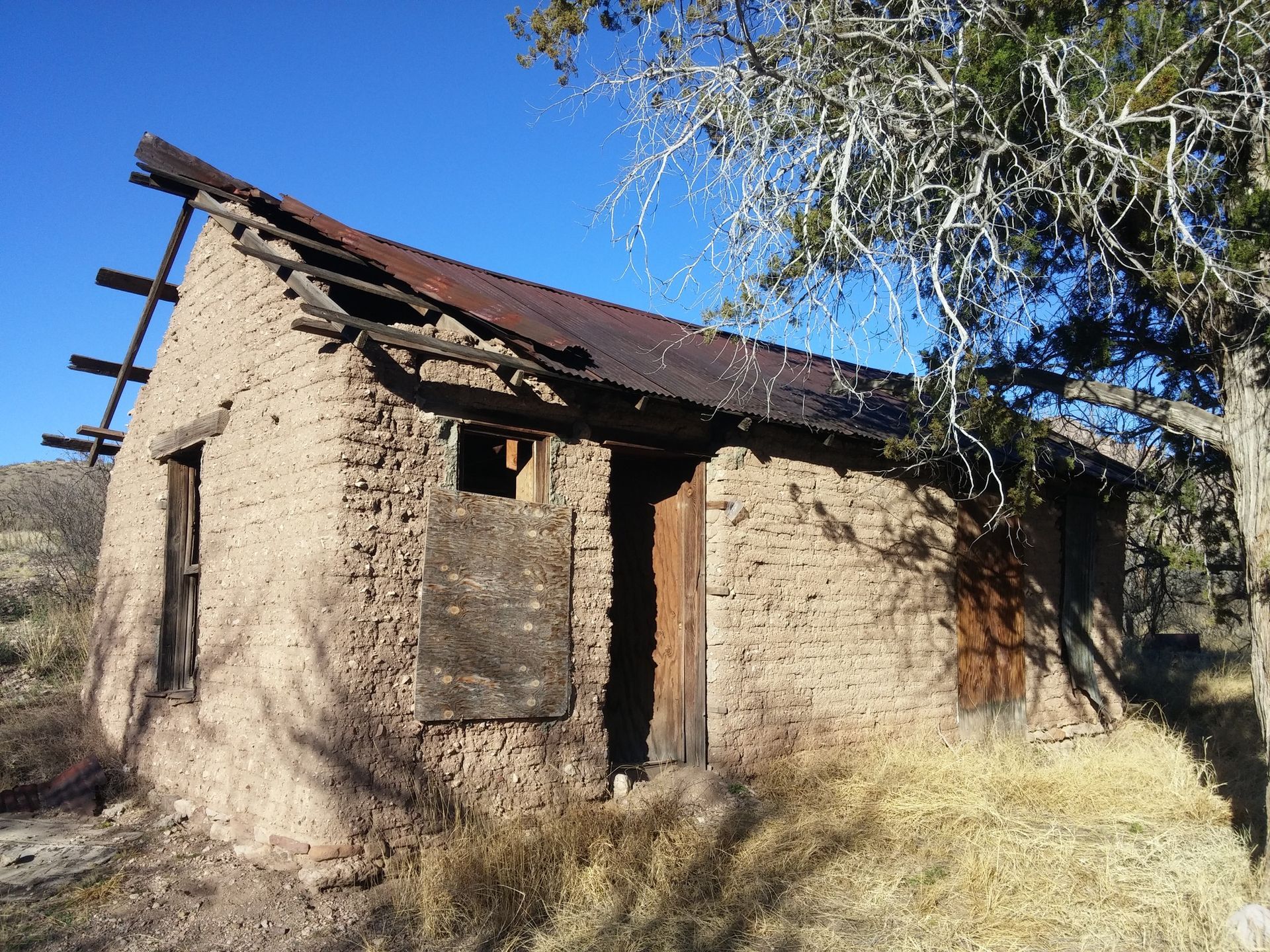 Dilapidated adobe structure with missing roof and boarded-up windows, set in a dry, outdoor landscape.