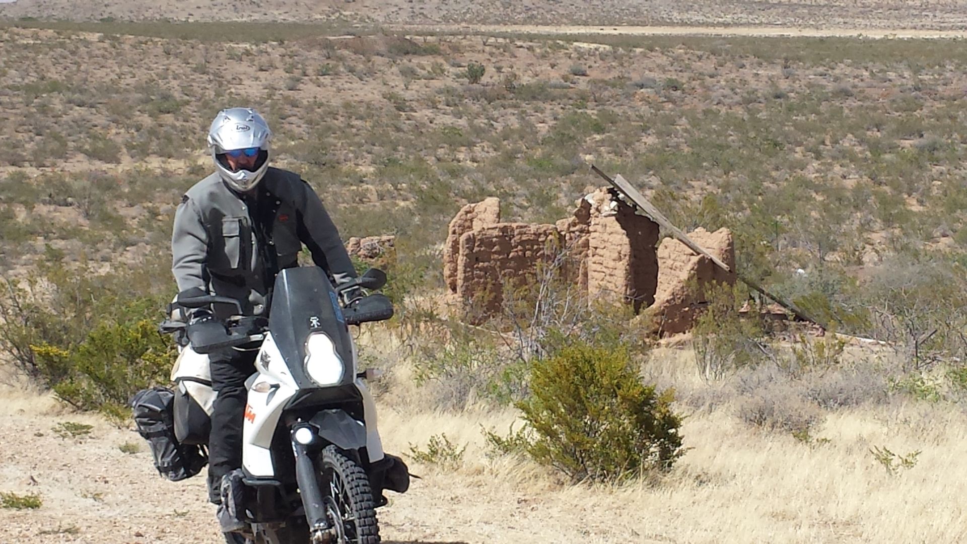 Motorcyclist on dirt road in arid landscape with old adobe building in the background.
