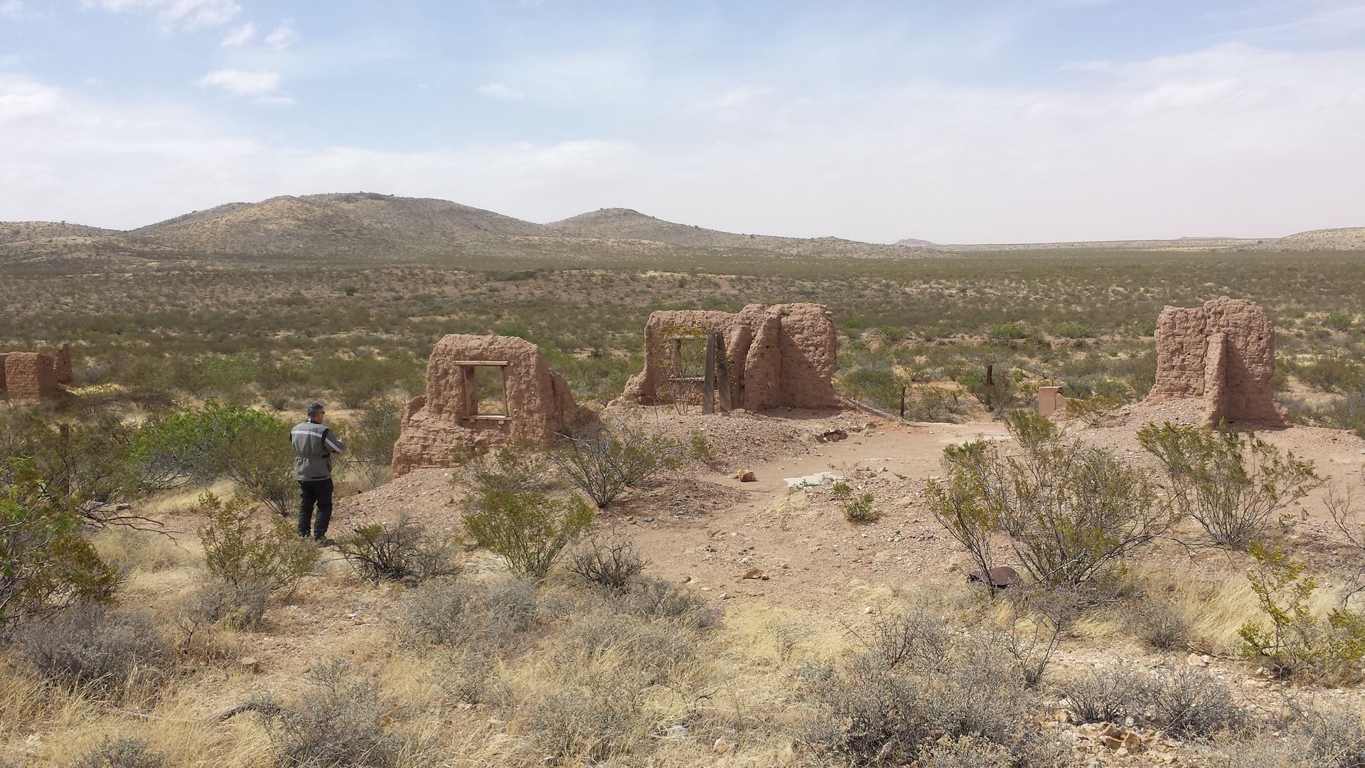 Ruined adobe structures in a desert landscape; a person stands nearby, looking toward the structures.