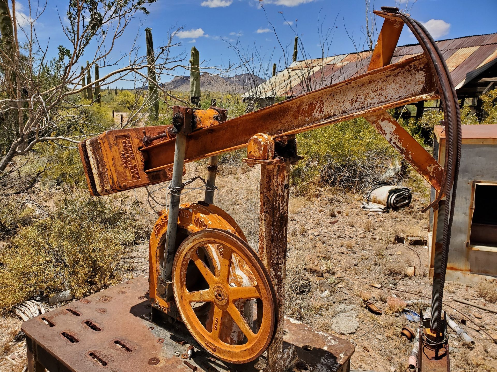 Rusty, orange mining pump in a desert setting with cacti and dilapidated buildings under a blue sky.