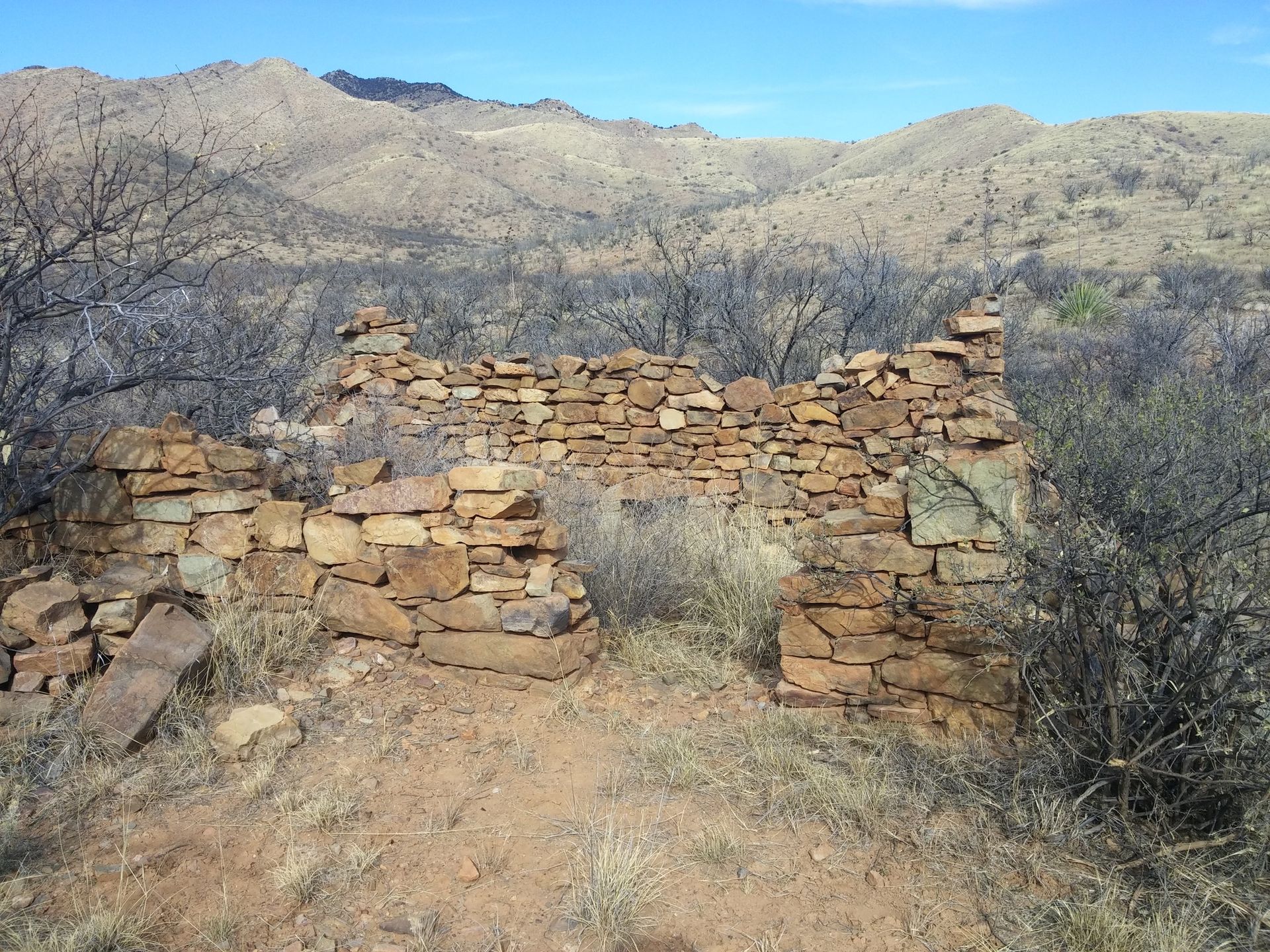 Stone ruins of a building in a desert landscape, with mountains in the background under a blue sky.