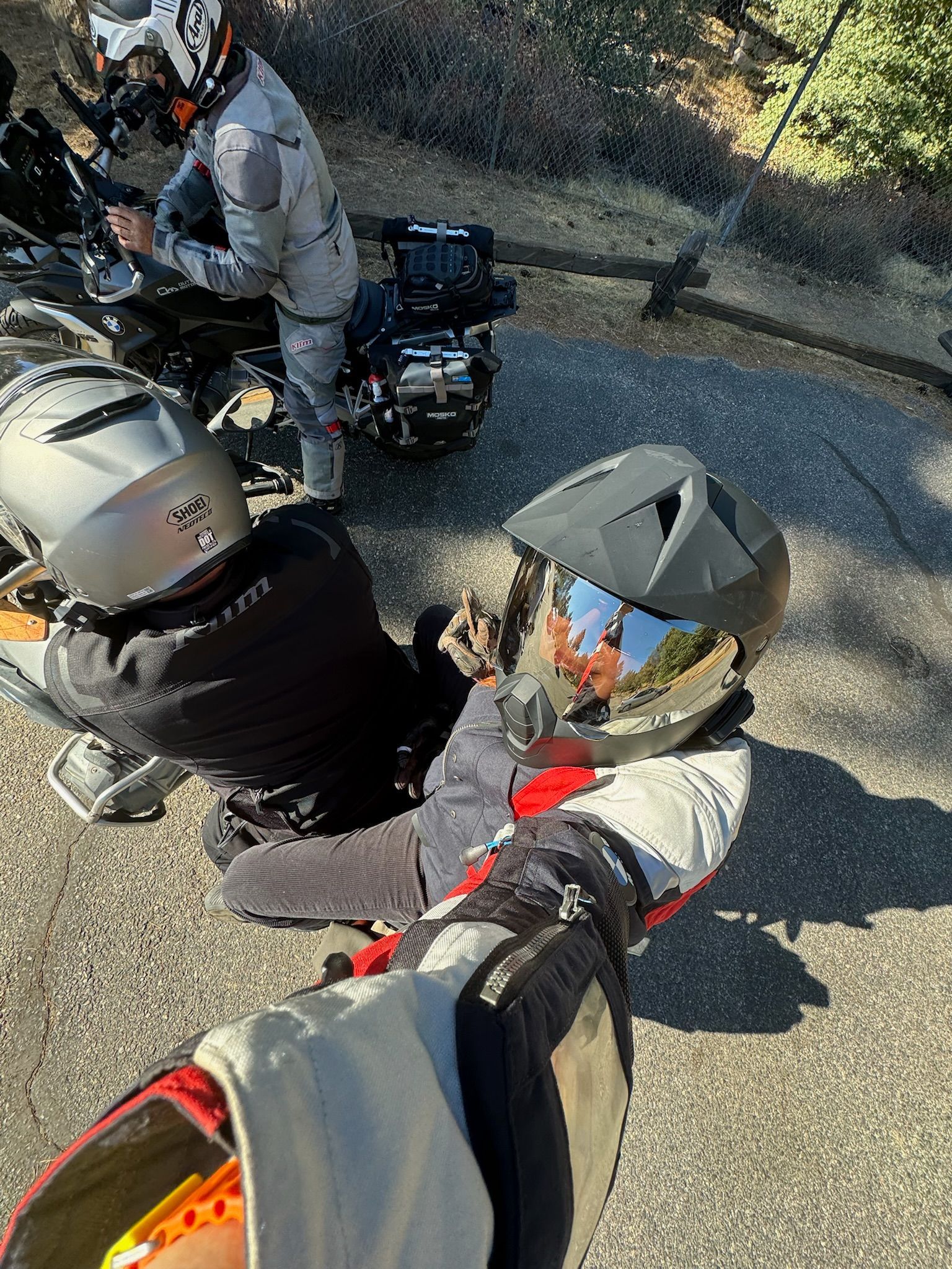Motorcyclists adjusting bikes on a roadside. Helmets, gear, asphalt, and natural setting visible.