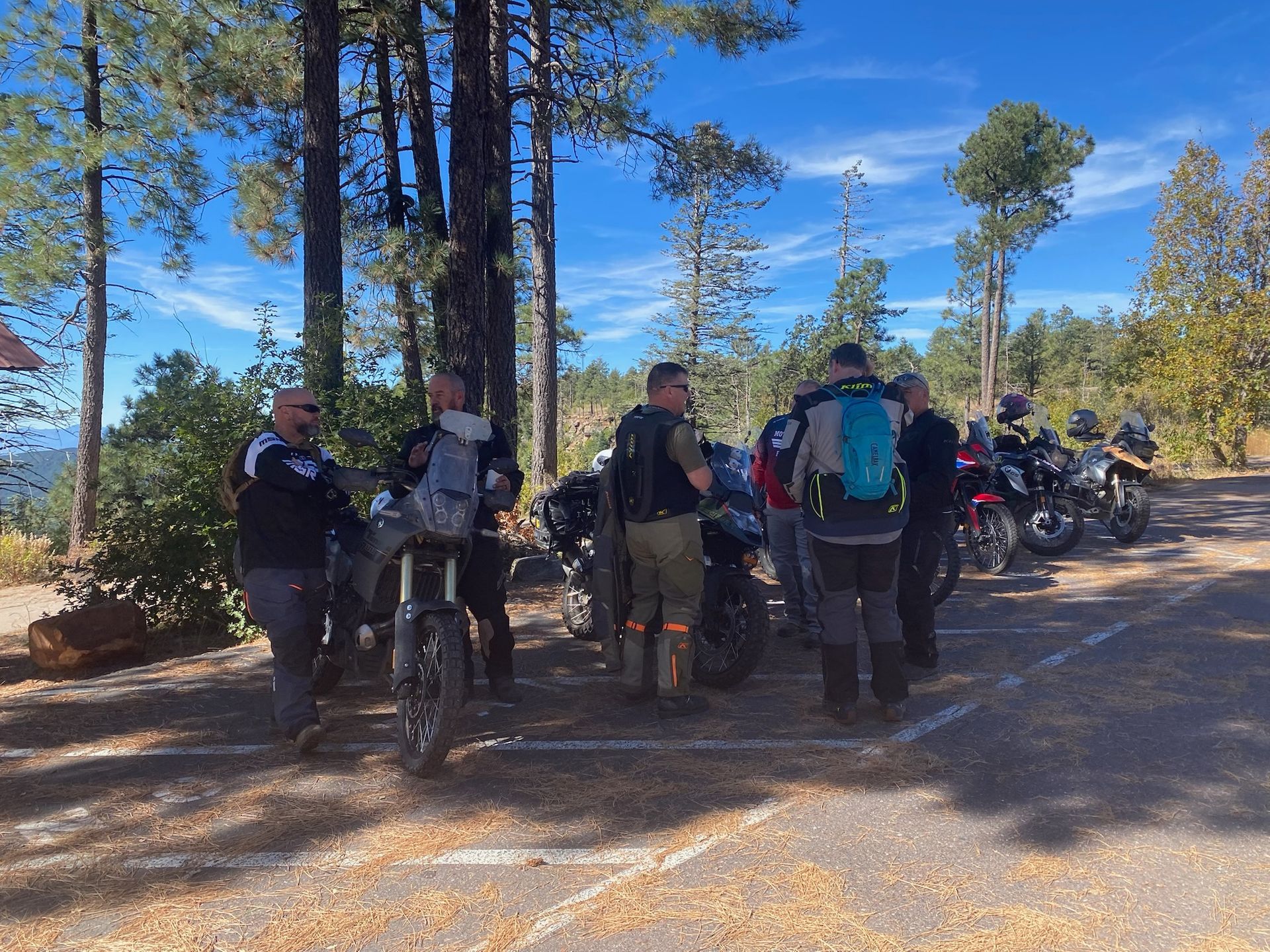 Group of motorcyclists standing near their bikes in a parking area, surrounded by trees under a blue sky.