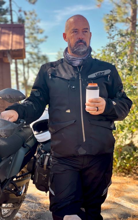 Man in motorcycle gear holding a drink, standing next to a motorcycle outdoors.