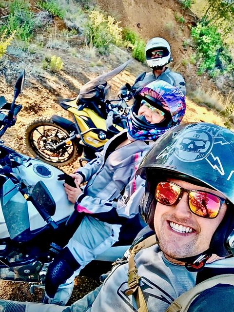 Three people on motorcycles on a dirt path, smiling. One in front wears a helmet and sunglasses, others in helmets.