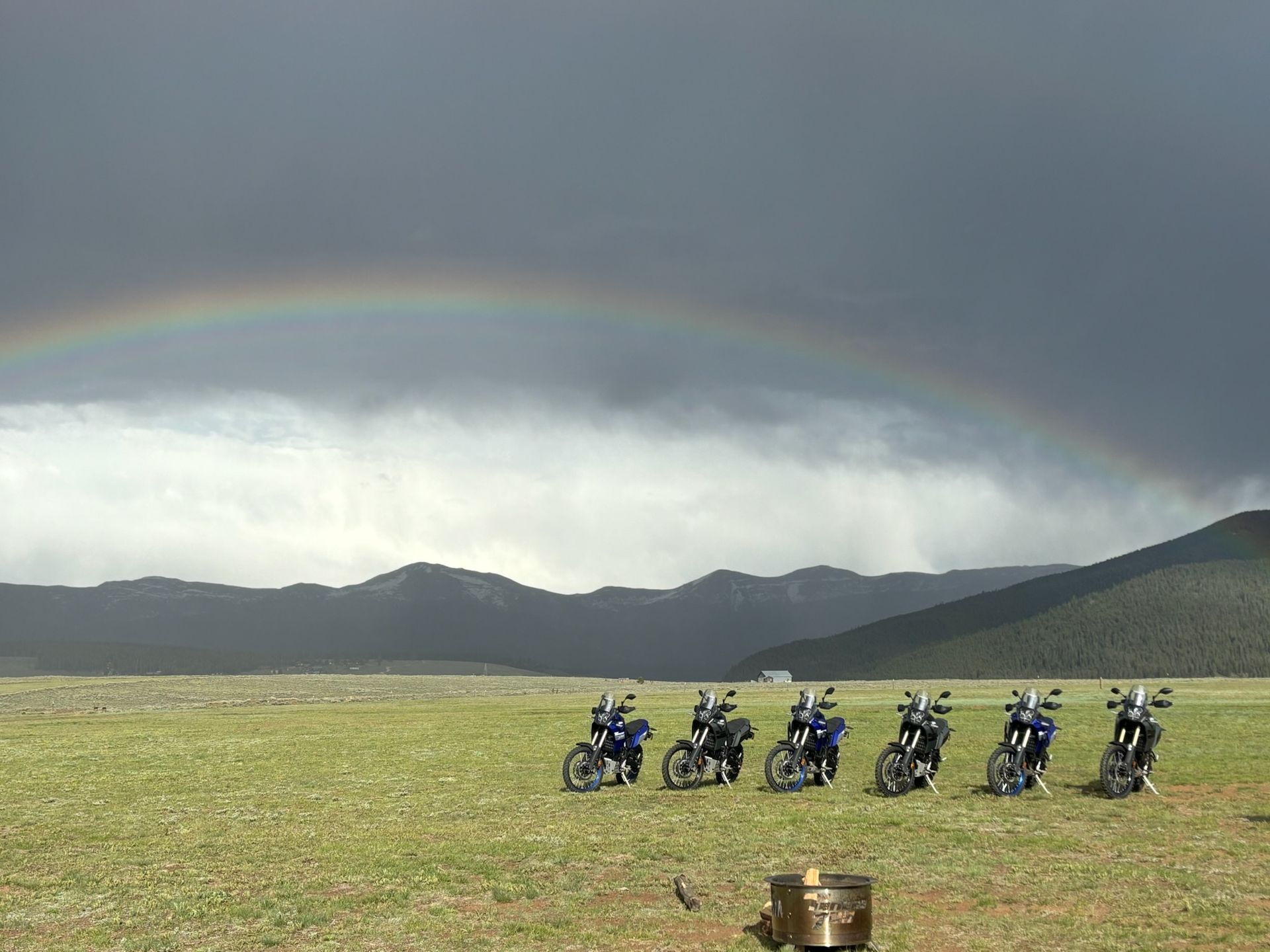 Motorcycles parked in a field with a rainbow arching overhead. Mountains and a dark sky are in the background.