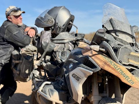 Man leaning on a dirty motorcycle in a desert setting.